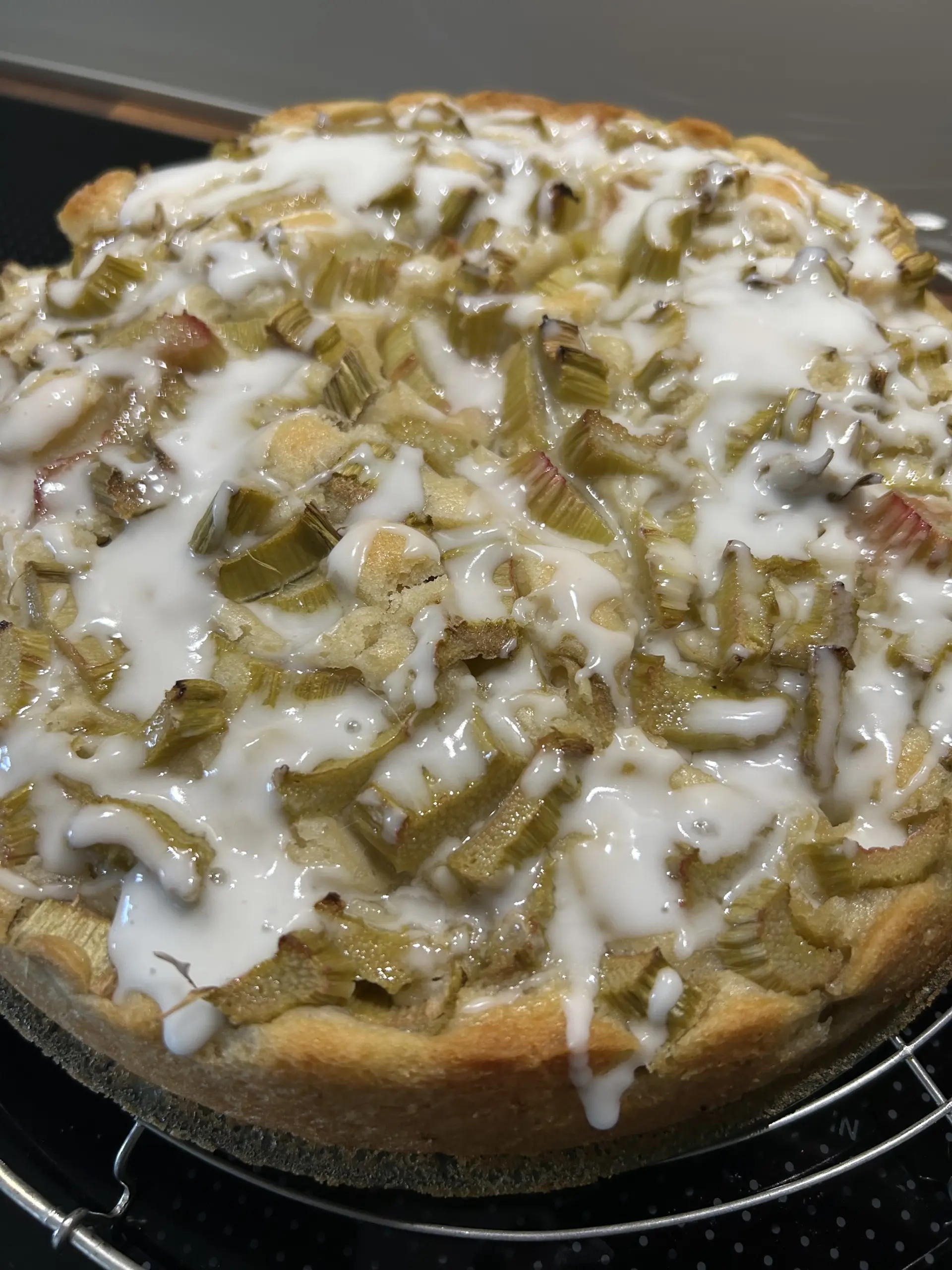 Ein runder Kuchen auf einem runden Kuchengitter. Oben sind teilweise versunkene Rhabarberstücke zu sehen. Darauf ungleichmäßig verteilter weißer Zuckerguss